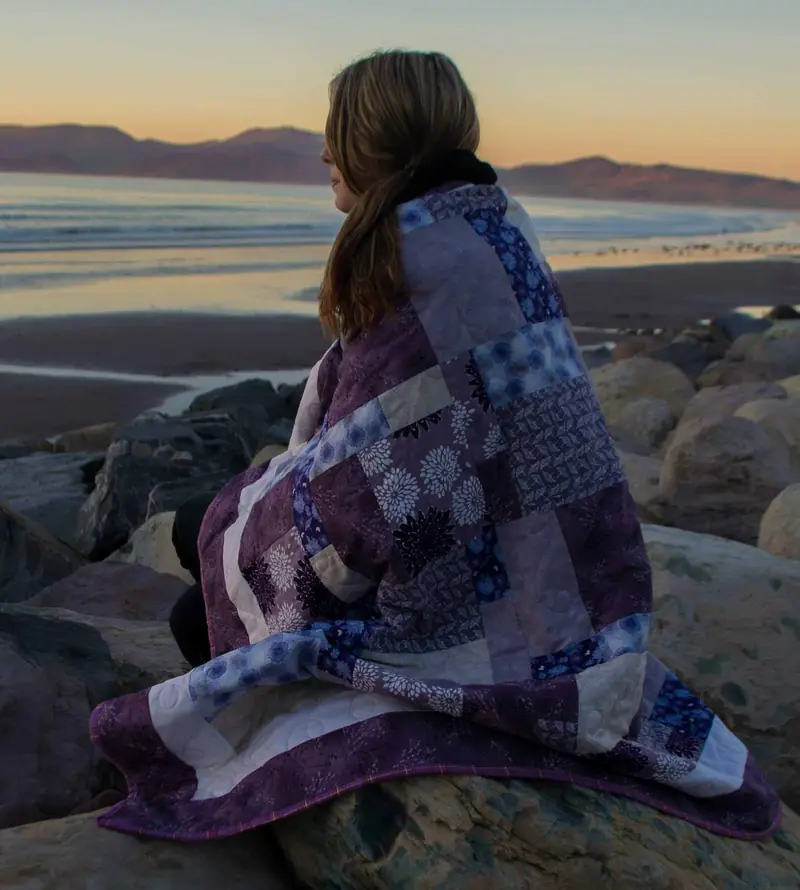 A woman wrapped in a handmade quilt watching the sunset by the sea